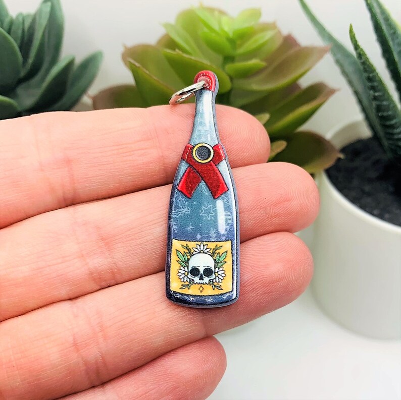 May include: A blue glass bottle charm with a skull and flowers on the label. The bottle has a red ribbon around the neck and a silver ring at the top.