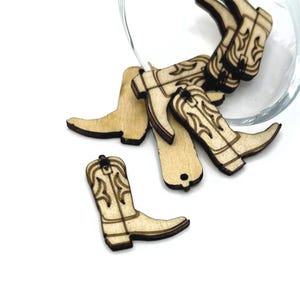 May include: A group of wooden cowboy boot charms in natural wood color. The boots have a detailed design and are ready to be used for crafting projects.