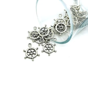 May include: A close-up of a glass jar filled with silver-toned metal charms shaped like ship steering wheels. The charms are scattered on a white surface.