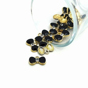 May include: A group of black and gold bow charms with rhinestones. The charms are scattered on a white surface and spilling out of a glass jar.
