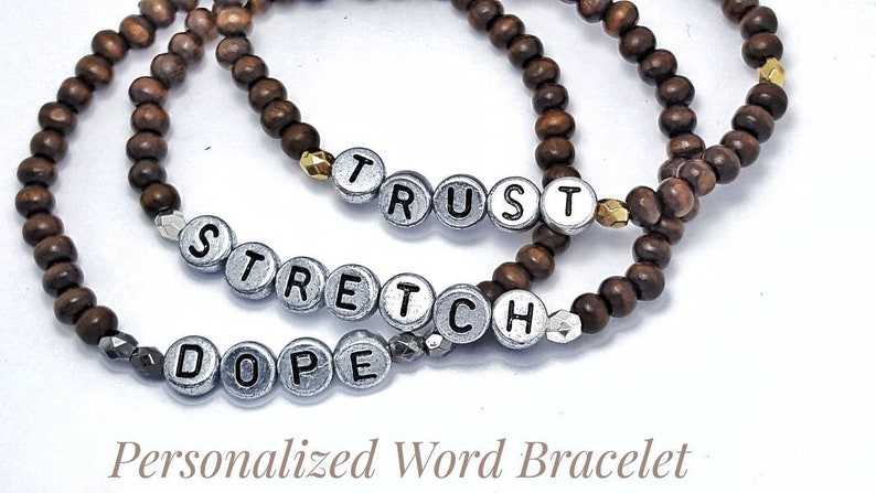 以下が含まれることがあります： 「Dope」、「Stretch」、「Trust」という言葉を綴る銀色の文字ビーズが付いた、茶色の木製ビーズブレスレット3本。