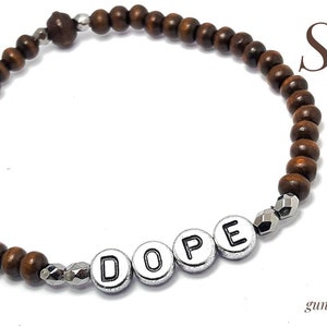 以下が含まれることがあります： 「DOPE」とスペルされたシルバーのアクセントビーズが付いたブラウンの木製ビーズブレスレット。ブレスレットは白い背景に映し出されています。ガンメタル、シルバー、またはゴールドのアクセントビーズをご用意しています。