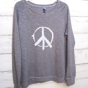 May include: A gray long-sleeved sweatshirt with a white peace sign graphic on the front. The sweatshirt has a crew neck and raglan sleeves. The peace sign is a simple, hand-drawn style.
