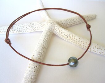 May include: A brown leather necklace with a single dark gray pearl. The necklace is knotted at both ends and rests on a white starfish. The pearl is approximately 0.3 inches in diameter.