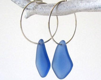 May include: Silver hoop earrings with light blue, teardrop-shaped glass pendants. The earrings hang from a white, textured branch. The pendants have a frosted finish. The background is white.