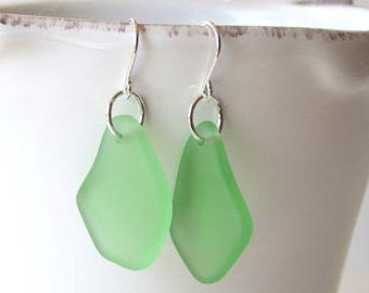 Pode incluir: Um par de brincos em forma de diamante verde claro com ganchos prateados. Os brincos têm uma aparência fosca de vidro do mar. Os ganchos são presos à parte superior dos brincos com pequenos anéis prateados. Fundo branco.