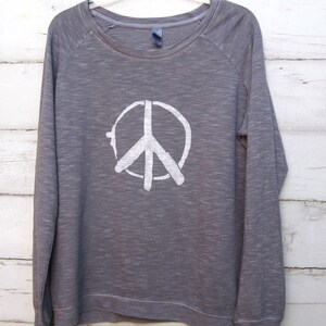 May include: A gray long-sleeved sweatshirt with a white peace sign graphic. The crew neck shirt has raglan sleeves and a relaxed fit. The peace symbol is centered on the front of the shirt.