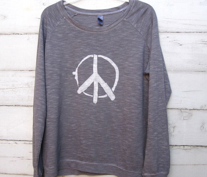 May include: A gray long-sleeved sweatshirt with a white peace sign graphic. The crew neck shirt has raglan sleeves and a relaxed fit. The peace symbol is centered on the front of the shirt.