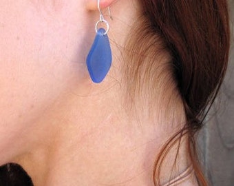 Könnte beinhalten: Ein einzelner blauer, tropfenförmiger Ohrring mit einem silbernen Haken. Der Ohrring hat einen leuchtenden Blauton und scheint aus Glas oder einem ähnlichen Material gefertigt zu sein. Der Ohrring hängt an einem silbernen Haken.