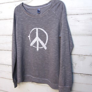 May include: A gray long-sleeved sweatshirt with a white peace sign graphic. The crew neck shirt is hanging on a white hanger against a white wooden background. The peace sign is a simple, bold design.