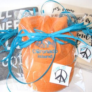 May include: Several gift items wrapped in clear plastic and tied with turquoise raffia ribbon. Items include a gray t-shirt with white text, an orange hat with blue embroidery, and a beige item with black script. Each item has a white tag with a peace sign.