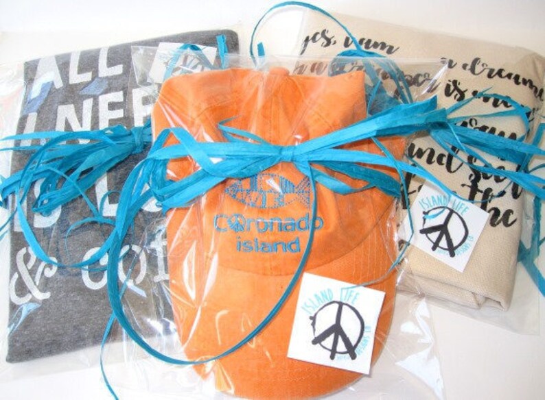 May include: Several gift items wrapped in clear plastic and tied with turquoise raffia ribbon. Items include a gray t-shirt with white text, an orange hat with blue embroidery, and a beige item with black script. Each item has a white tag with a peace sign.