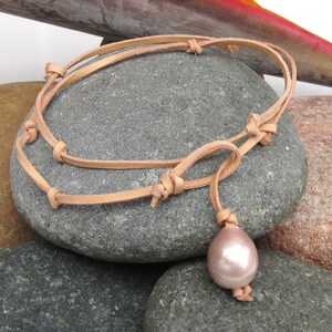 Könnte beinhalten: Ein hellbraunes Lederarmband mit Knotenmuster und einem rosa, tropfenförmigen Perlenanhänger. Das Armband wird auf einem grauen Stein präsentiert, der die natürlichen Materialien und die neutrale Farbpalette hervorhebt.