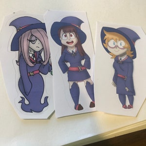 May include: Three cartoon witch stickers featuring a purple witch with a star on her hat, a brown-haired witch with a red bow, and a blonde witch with glasses. The witches are wearing blue and black outfits and are standing on white backgrounds.