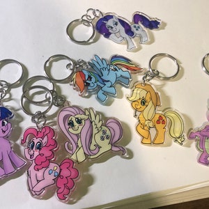 May include: A set of eight My Little Pony keyrings featuring different characters, including Twilight Sparkle, Pinkie Pie, Fluttershy, Rainbow Dash, Rarity, Applejack, Spike, and a unicorn. The keyrings are made of clear acrylic and feature colourful illustrations of the characters.