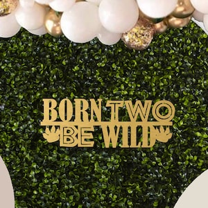 May include: A wooden table with a white cake, cookies, and cupcakes on it. The table is decorated with a green leafy backdrop and a gold sign that says "Born Two Be Wild".