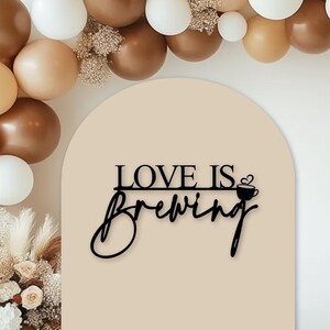 May include: A beige arch with the text "LOVE IS BREWING" in black with a small coffee cup icon. The arch is decorated with a garland of brown and white balloons.