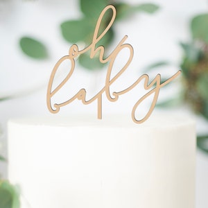 May include: A white frosted cake with a wooden "oh baby" cake topper. The cake sits on a white cake stand. Green foliage and a pink vase with flowers are in the background, creating a soft, celebratory scene.