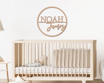 Może przedstawiać: Drewniany znak z imieniem "NOAH James" w okrągłym wzorze, zamontowany nad łóżeczkiem. Znak jest wykonany z jasnego drewna, a pokój dziecięcy ma neutralną kolorystykę.