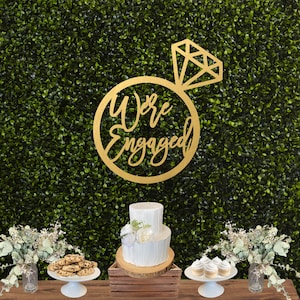 May include: A gold wooden sign with the words "We're Engaged" in a circle with a diamond ring on top. The sign is hanging on a green wall with a white cake and cupcakes on a wooden table in front of it.