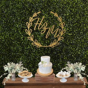 May include: A wooden table displays a two-tiered white cake with floral accents, cookies, and cupcakes. A gold-colored monogram with the letters "H" and "M" is set against a green hedge backdrop. Decorative floral arrangements are in glass jars.