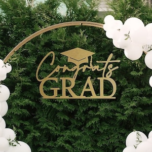 May include: A graduation party backdrop with a gold archway decorated with white and light blue balloons. The archway has a sign that says "Congrats Grad" with a graduation cap.