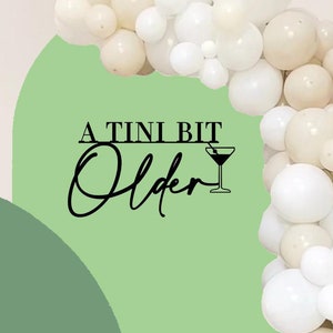 May include: A green and white balloon arch with the text "A TINI BIT Older" and a martini glass icon. The balloons are white and cream.