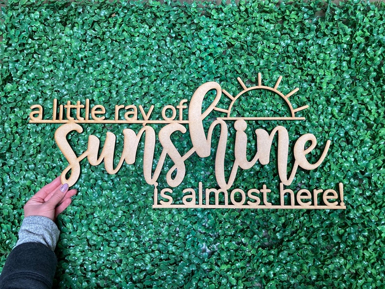 May include: A wooden sign with the text "a little ray of sunshine is almost here!" in a cursive font. The sign also features a stylized sun with rays.