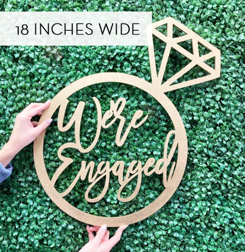 May include: A wooden ring with the words "We're Engaged" in cursive script. The ring is topped with a diamond shape. The sign is 18 inches wide.