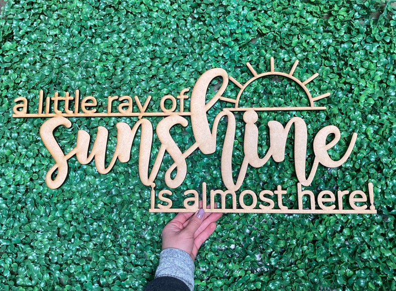 May include: A wooden sign with the text "a little ray of sunshine is almost here!" The sign features a stylized sun graphic.