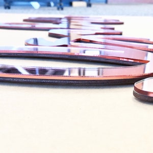 May include: Rose gold mirrored acrylic word art. The letters are cut from a reflective material with a dark brown border. The word is lying on a light tan surface. The letters are approximately 1 inch thick.