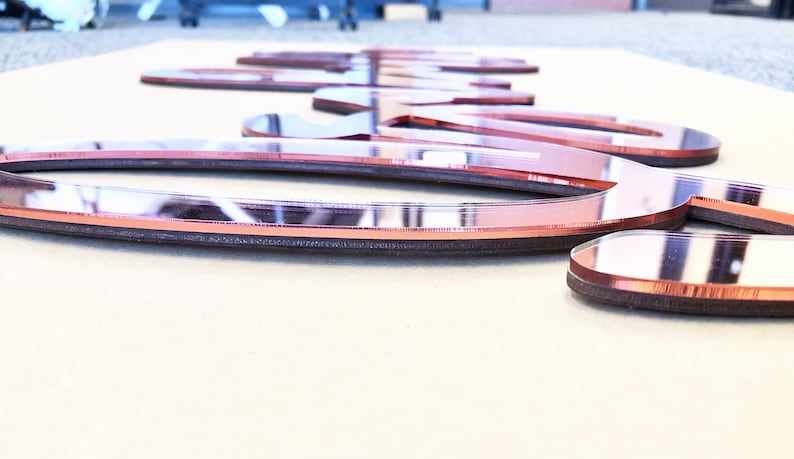 May include: Rose gold mirrored acrylic word art. The letters are cut from a reflective material with a dark brown border. The word is lying on a light tan surface. The letters are approximately 1 inch thick.