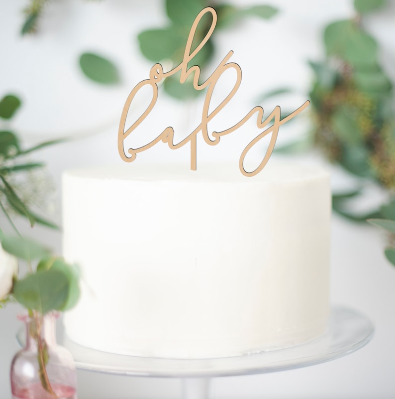 May include: A white frosted cake with a wooden "oh baby" cake topper. The cake sits on a white cake stand. Green foliage and a pink vase with flowers are in the background, creating a soft, celebratory scene.