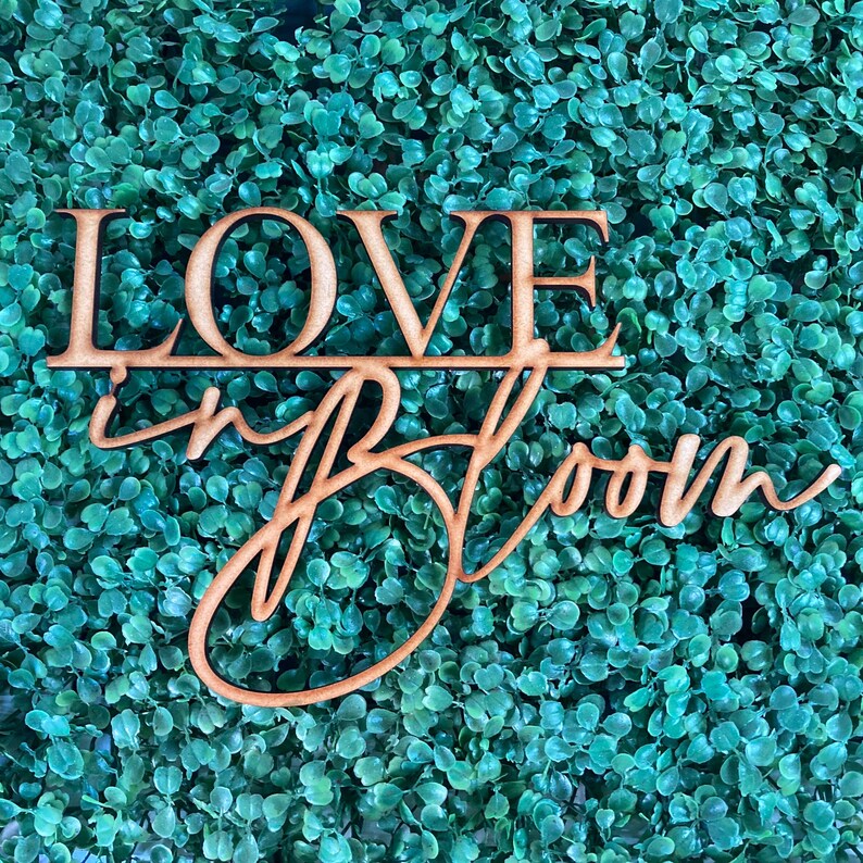 Op de afbeelding: Een houten bord met de tekst "LOVE in BLOOM" in een cursief schrift. Het bord is bruin en is geplaatst tegen een achtergrond van groen kunstmatig gebladerte.
