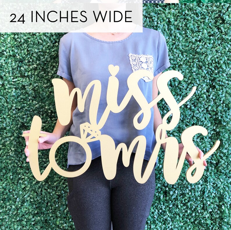 May include: Gold wooden sign with the words "miss to mrs" in a cursive font. The sign is 24 inches wide and features a ring with a diamond.