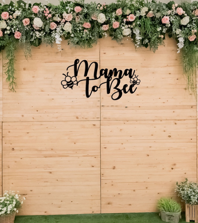 May include: A wooden backdrop with the phrase "Mama to Bee" in black script, adorned with bee illustrations. The backdrop is decorated with floral arrangements of pink and white roses, greenery, and hanging vines, suitable for a baby shower.
