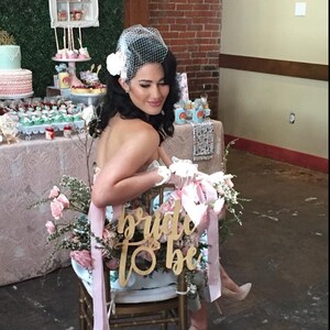 May include: A bride-to-be sits on a chair with a pink and white floral decoration and a wooden sign that says "bride to be".