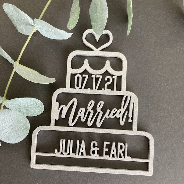 May include: A silver-colored cake-shaped wedding ornament with the date "07.17.21", the word "Married!", and the names "JULIA & EARL" cut out. A heart shape tops the cake design. The ornament is set against a dark background with green foliage.