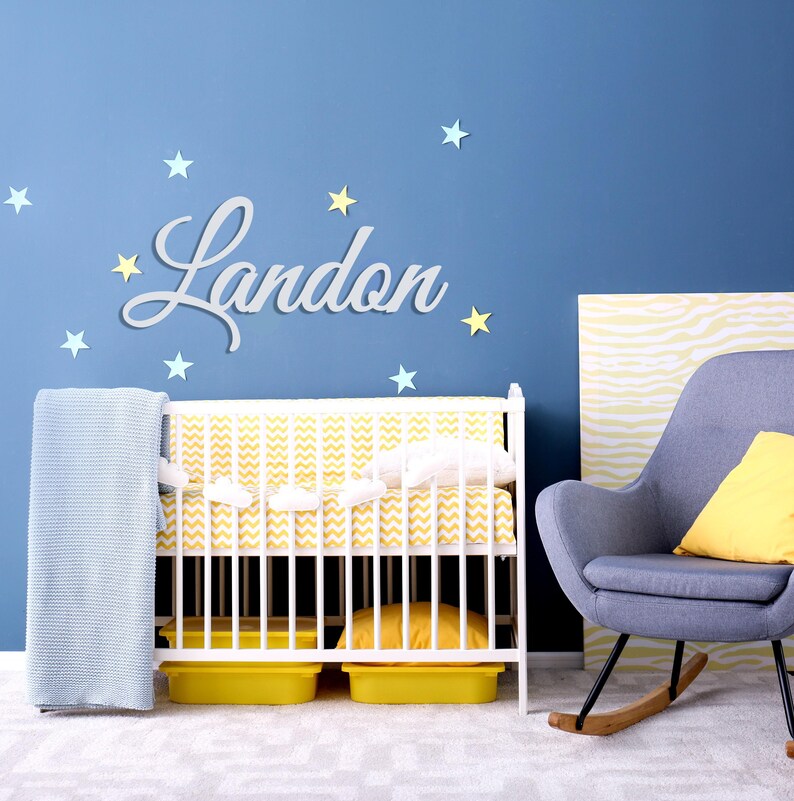 May include: A nursery scene with a white crib, a blue wall, and the name "Landon" in cursive script. The crib has yellow and white bedding, a blue blanket, and yellow storage bins. Decorative stars are scattered around the name.