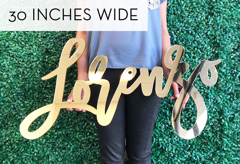 May include: A gold-colored, cursive-style sign that reads "Lorenzo" is held in front of a green hedge. The sign is 30 inches wide. The letters are connected and have a reflective, mirror-like finish.