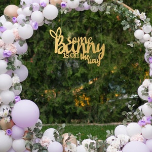 May include: A gold sign with the text "some bunny is on the way" hanging from a circular archway decorated with white and lavender balloons, greenery, and white flowers.
