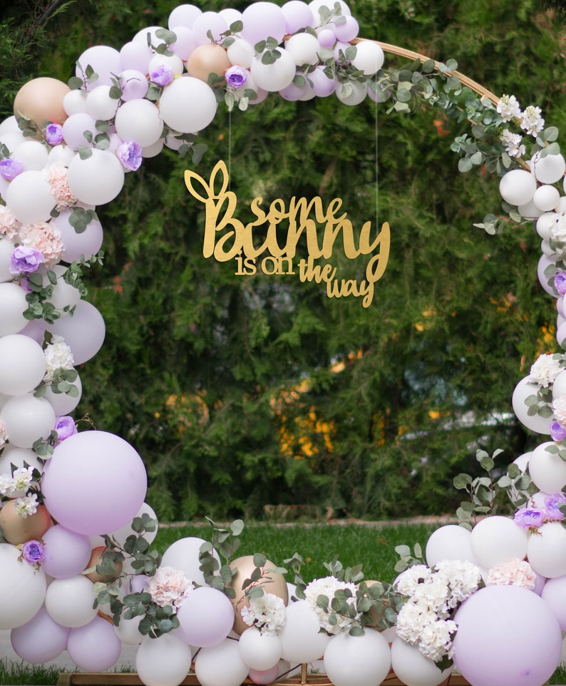 May include: A gold sign with the text "some bunny is on the way" hanging from a circular archway decorated with white and lavender balloons, greenery, and white flowers.