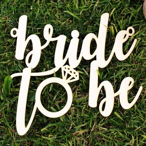 May include: Wooden sign with the words "bride to be" in a cursive font. The sign is decorated with a ring and diamond.