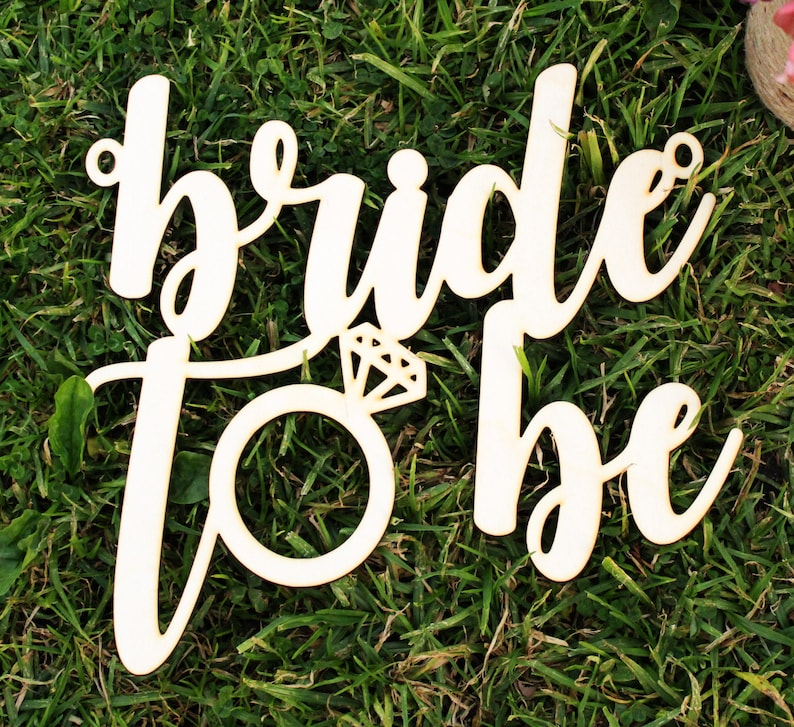 May include: Wooden sign with the words "bride to be" in a cursive font. The sign is decorated with a ring and diamond.