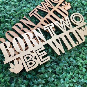 May include: Wooden sign with the text "Born Two Be Wild" in a rustic style. The sign is made of two pieces of wood, one with the text "Born Two" and the other with the text "Be Wild".