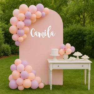 May include: A pink arch backdrop with the name "Camila" in white script, decorated with pastel balloons. A white table holds a cake and desserts. The scene is set outdoors on a green lawn.