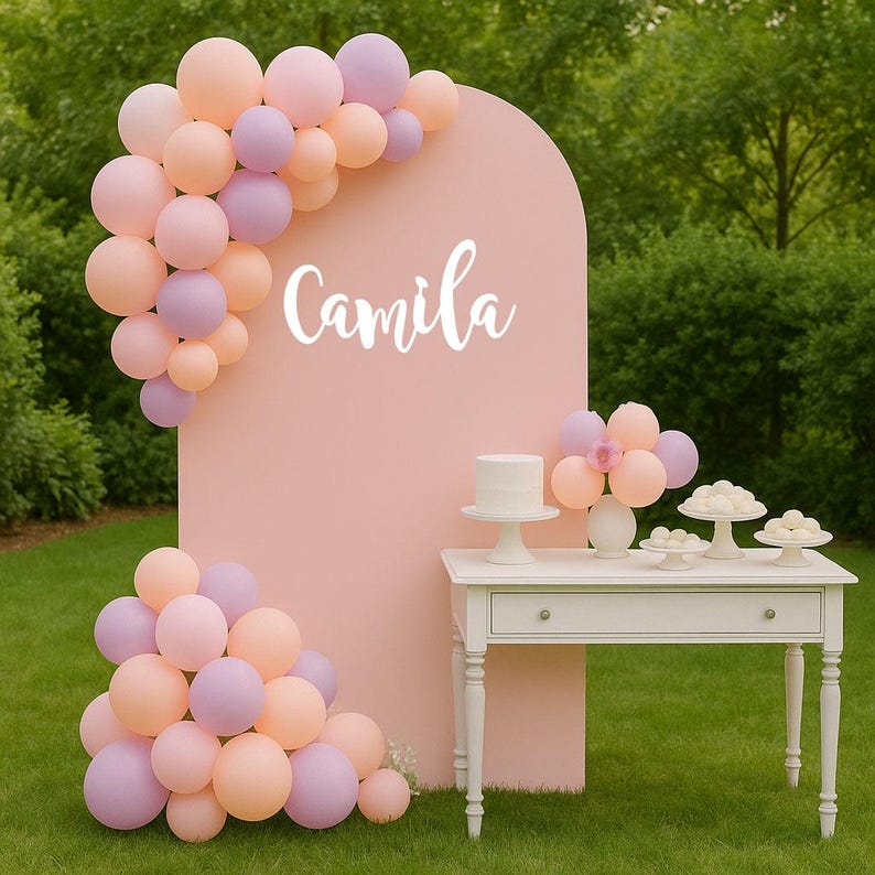 May include: A pink arch backdrop with the name "Camila" in white script, decorated with pastel balloons. A white table holds a cake and desserts. The scene is set outdoors on a green lawn.