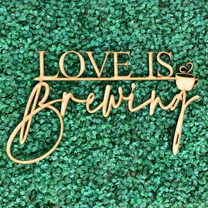 May include: Wooden sign with the text "Love is brewing" in cursive script. A small coffee cup with a heart is above the word "brewing".