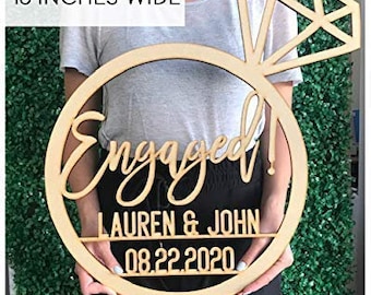 May include: Wooden ring-shaped sign with the word "Engaged!" in cursive script, the names "Lauren & John" and the date "08.22.2020". A diamond ring shape is attached to the top of the sign. The sign is 18 inches wide.
