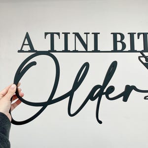 May include: A black metal sign with the text "A TINI BIT Older" in a cursive font. A martini glass with a straw is to the right of the text.